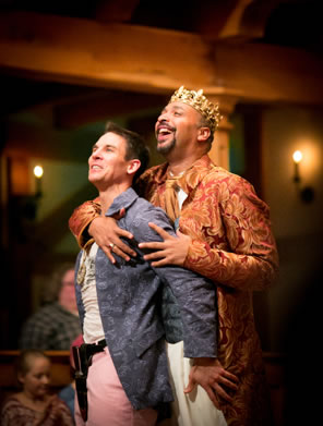 Gaveston has his back pressed up against Edward, and left hand on the king's thigh, as Edward puts his arms around Gaveston's chest, hands on the left shoulder, and both are smiling with delight: Blackfriars Playhouse candles and audience members visible in the wood-beamed background.